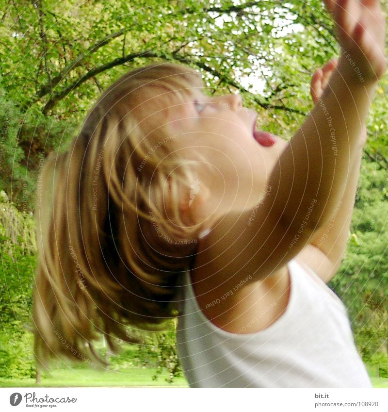 Lustiges, witziges, fröhliches, glückliches, ulkiges, blondes Mädchen draussen im Garten, schaut nach oben und streckt dabei die beiden Arme hoch zum Baum. Kleiner Spaßvogel macht Quatsch, streckt die Zunge raus, in der Natur, im Park unter dem Baum.