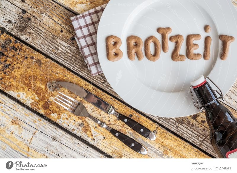 Auf einem rustikalen Holztisch ein Teller mit den Buchstaben BROTZEIT, Messer und Gabel, Serviette und eine Flasche Bier im Querformat Lebensmittel Brot