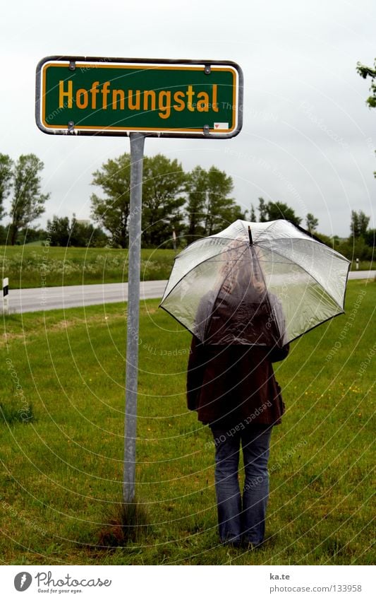 es gibt HOFFNUNG Hoffnung Regenschirm Ortsschild grün Außenaufnahme Frau durchsichtig Gras Straßenrand Baum graue Wolken trist nass Trauer Götter stehen