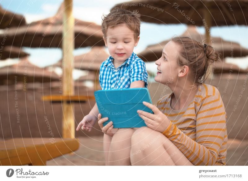 Kleiner Junge mit seiner Mutter in einem Strandresort spielen mit einem Tablet-Computer am Strand unter Stroh Sonnenschirme Freude Glück Spielen