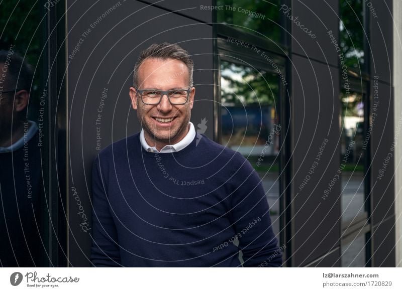 Gut aussehender, lächelnder Mann mittleren Alters mit Brille und Pullover, der sich an ein Gebäude lehnt, in dem sich die Bäume im Fenster spiegeln Glück