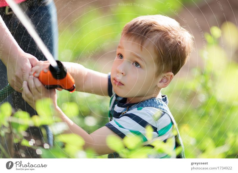 Niedlicher ernster kleiner Junge, der mit einem Wasserstrahl aus einem Sprinkler spielt, der von seiner Mutter im Garten gehalten wird, während er zwischen grünen, belaubten Sträuchern steht