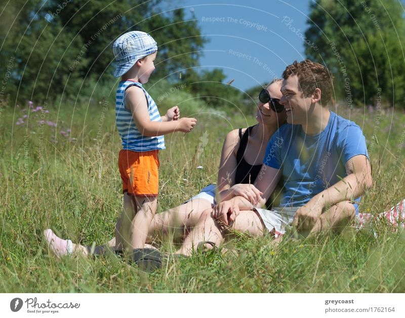 Junge Eltern und Sohn auf der Wiese an einem heißen Sommertag. Glückliches Paar sitzt auf dem Gras und schaut das Kind an. Outdoor-Familienaktivität Freude