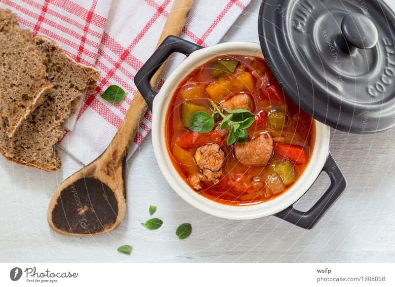 Gulasch in einer schwarzen cocotte mit Majoran auf weißem Holz Brot Suppe Eintopf Kochlöffel dunkel Goulasch Gulyas Gollasch Gujasch Ungarisch essen Paprika