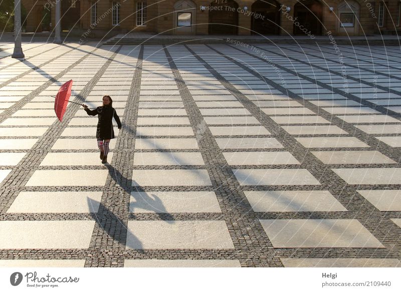 Rückansicht einer Frau mit dunklem Mantel und Regenschirm auf einem großen gepflasterten Platz im Gegenlicht mit Schatten Mensch feminin Erwachsene 1