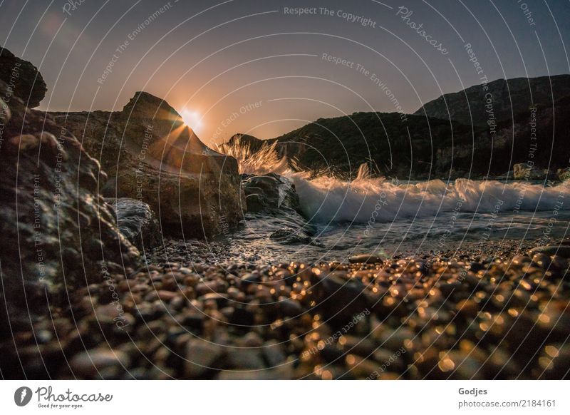 Liapades Beach, Abendstimmung an einem steinigen Strand, Wellen brechen an einem Felsen hinter dem die Sonne untergeht Umwelt Wasser Wolkenloser Himmel