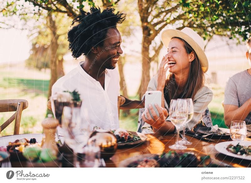 Gemischte Rennpaare, die Spaß mit dem Telefon bei der Dinnerparty haben. Mittagessen Abendessen Alkohol Freude Glück Sommer Sonne Restaurant Feste & Feiern