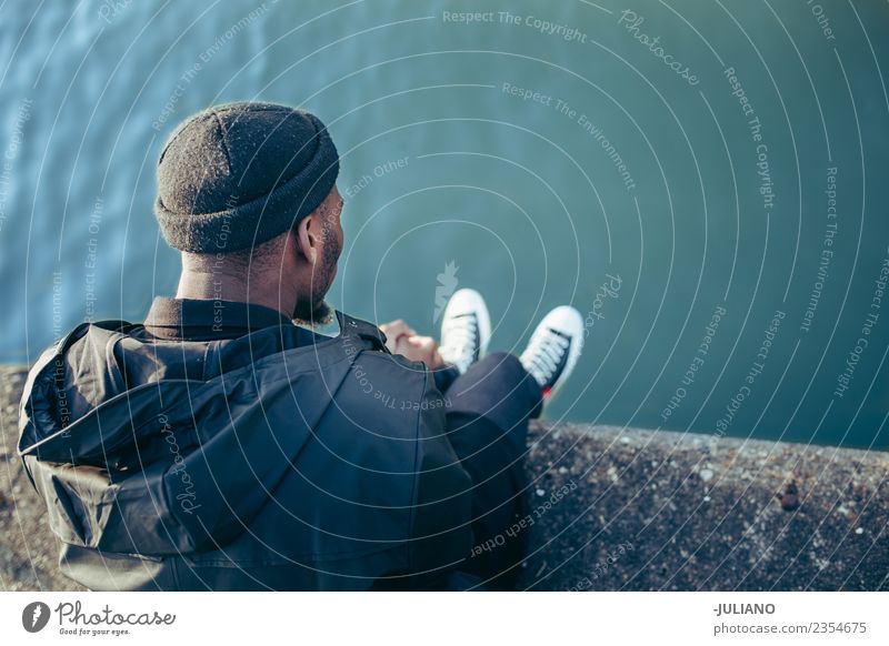 Junger Mann, der am Hafen sitzt und auf das Wasser schaut. Lifestyle Mensch Jugendliche 13-18 Jahre 18-30 Jahre Erwachsene Frühling schlechtes Wetter Wind Hut