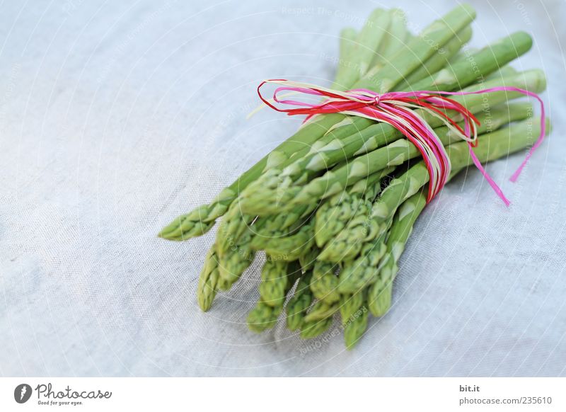 Eine Portion grüner Spargel, frisch geerntet vom einheimischen Feld, liegt dekoriert mit einem kariertem Band kariertem aus Stoff, zusammen gebunden, im Licht auf dem Tisch, auf einem weißem Tuch aus Leinen.