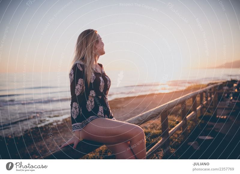 Junge Frau, die auf einem Geländer sitzt und den Sonnenuntergang am Meer beobachtet. Strand blond Coolness Abenddämmerung Freude Mädchen gut Güte lachen