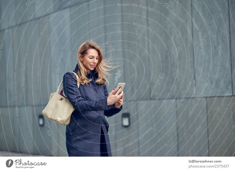 Blonde Frau mittleren Alters, die im Wind geht. Gesicht lesen Telefon Erwachsene 1 Mensch 45-60 Jahre Frühling Mantel blond Beton Bewegung modern selbstbewußt