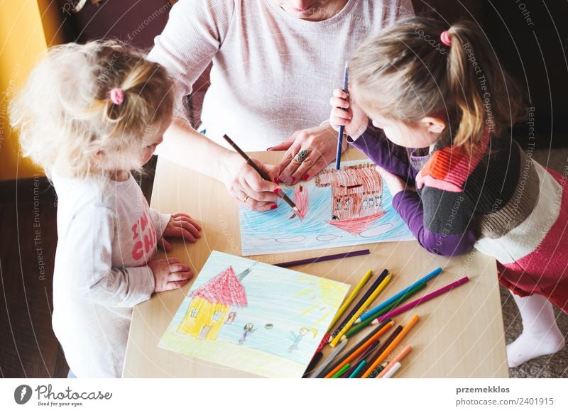 Mutter mit kleinen Mädchen, die ein buntes Bild von Haus und spielenden Kindern mit Bleistiftmalen malen, die drinnen am Tisch stehen Lifestyle Freude Glück