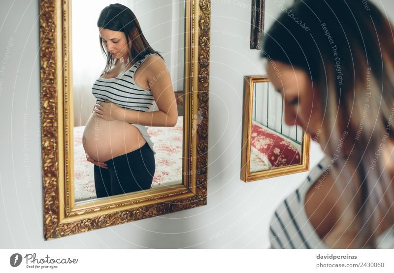 Schwangere Frau, die ihren Bauch ansieht. Lifestyle schön Leben Spiegel Schlafzimmer Mensch Baby Erwachsene Eltern Mutter Hand Denken Liebe Wachstum warten