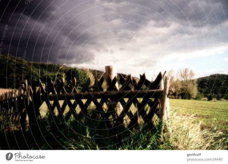 Jägerzaun Zaun Holz Holzzaun Wolken Regen dunkle Wolken Wiese Gras Baum dunkel grün gelb Gartenzaun Stimmung jägerzaun Himmel Landschaft hell blau