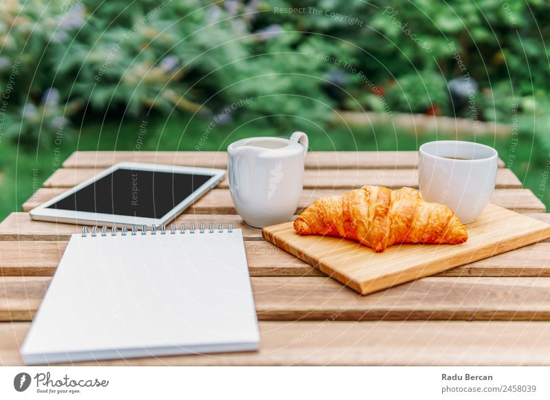 Frühstück am Morgen im Grünen Garten mit französischem Croissant, Kaffeetasse, Orangensaft, Tablette und Notizbuch auf Holztisch Tisch Hintergrundbild Sommer