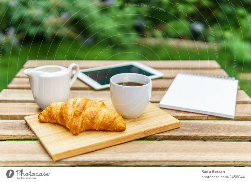 Frühstück am Morgen im Grünen Garten mit französischem Croissant, Kaffeetasse, Orangensaft, Tablette und Notizbuch auf Holztisch Tisch Hintergrundbild Sommer