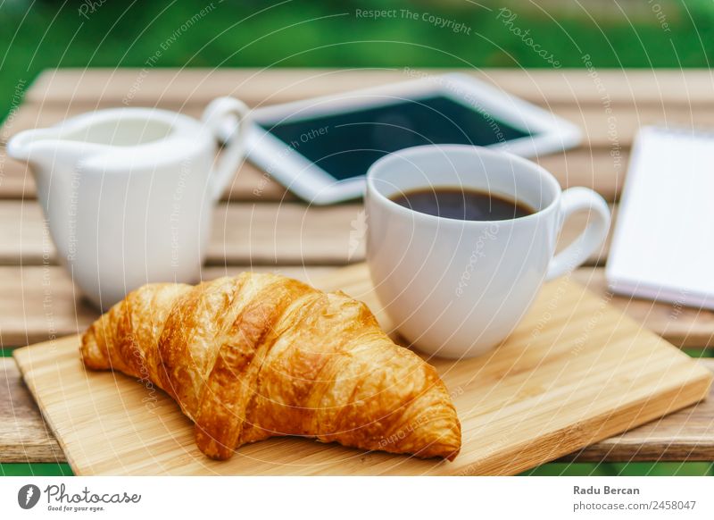 Frühstück am Morgen im Grünen Garten mit französischem Croissant, Kaffeetasse, Orangensaft, Tablette und Notizbuch auf Holztisch Tisch Hintergrundbild Sommer