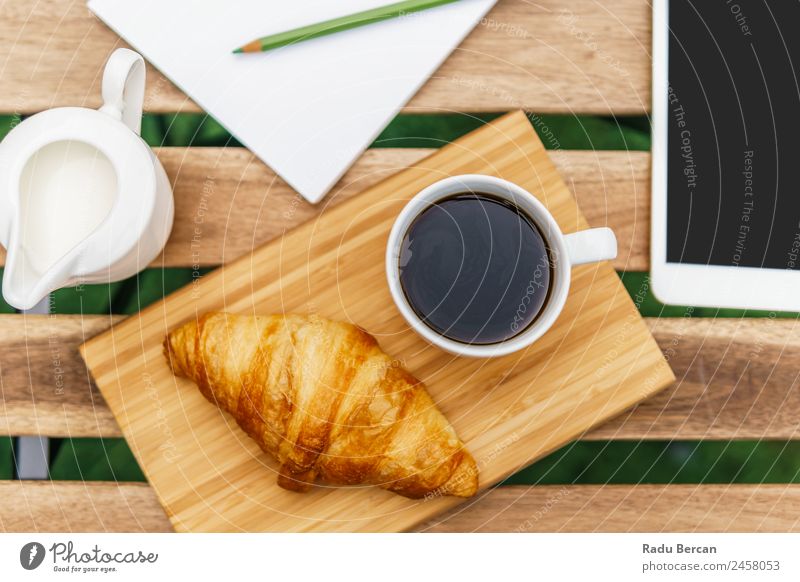 Frühstück am Morgen im Grünen Garten mit französischem Croissant, Kaffeetasse, Orangensaft, Tablette und Notizbuch auf Holztisch Tisch Hintergrundbild Sommer