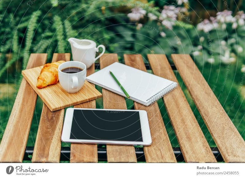 Frühstück am Morgen im Grünen Garten mit französischem Croissant, Kaffeetasse, Orangensaft, Tablette und Notizbuch auf Holztisch Tisch Hintergrundbild Sommer