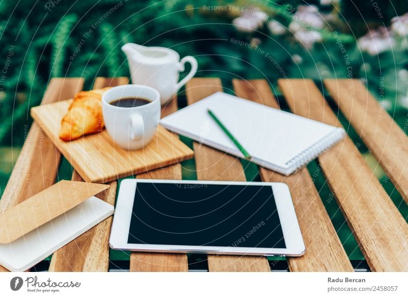 Frühstück am Morgen im Grünen Garten mit französischem Croissant, Kaffeetasse, Orangensaft, Tablette und Notizbuch auf Holztisch Tisch Hintergrundbild Sommer