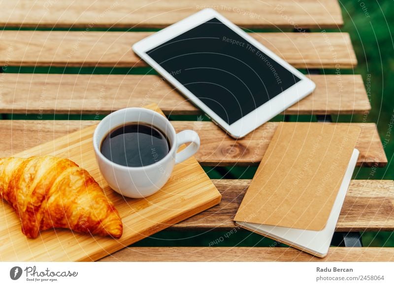 Frühstück am Morgen im Grünen Garten mit französischem Croissant, Kaffeetasse, Orangensaft, Tablette und Notizbuch auf Holztisch Tisch Hintergrundbild Sommer