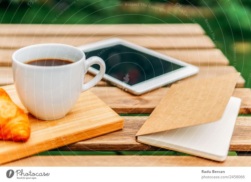 Frühstück am Morgen im Grünen Garten mit französischem Croissant, Kaffeetasse, Orangensaft, Tablette und Notizbuch auf Holztisch Tisch Hintergrundbild Sommer