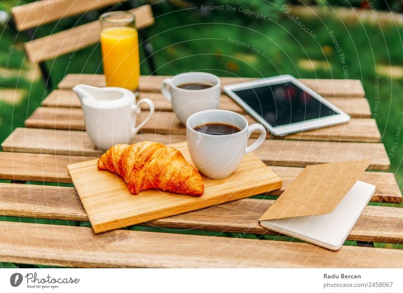 Frühstück am Morgen im Grünen Garten mit französischem Croissant, Kaffeetasse, Orangensaft, Tablette und Notizbuch auf Holztisch Tisch Hintergrundbild Sommer