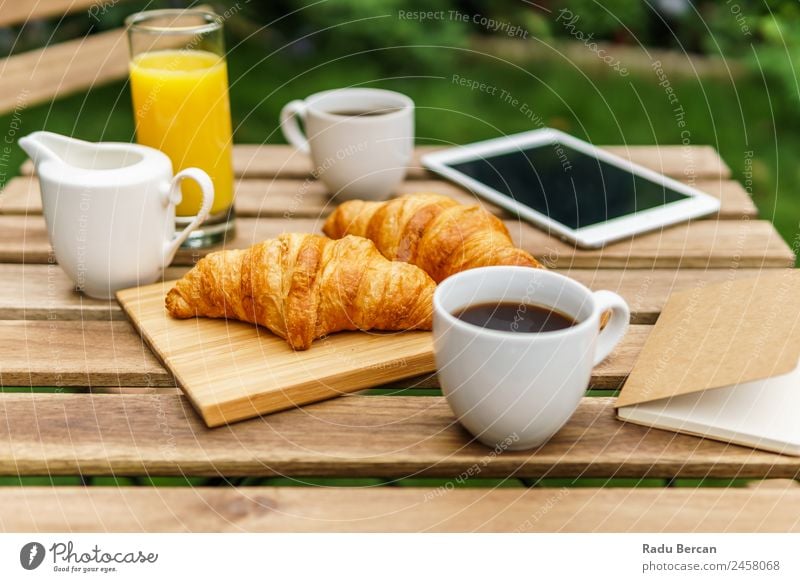 Frühstück am Morgen im Grünen Garten mit französischem Croissant, Kaffeetasse, Orangensaft, Tablette und Notizbuch auf Holztisch Tisch Hintergrundbild Sommer