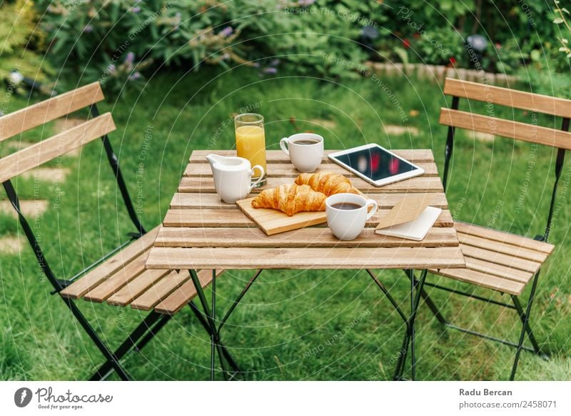 Frühstück am Morgen im Grünen Garten mit französischem Croissant, Kaffeetasse, Orangensaft, Tablette und Notizbuch auf Holztisch Tisch Hintergrundbild Sommer