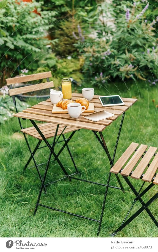 Frühstück am Morgen im Grünen Garten mit französischem Croissant, Kaffeetasse, Orangensaft, Tablette und Notizbuch auf Holztisch Tisch Hintergrundbild Sommer