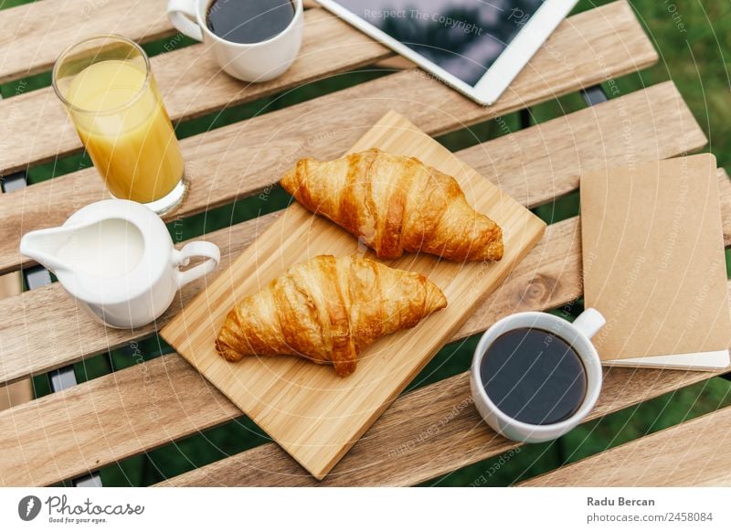 Frühstück am Morgen im Grünen Garten mit französischem Croissant, Kaffeetasse, Orangensaft, Tablette und Notizbuch auf Holztisch Tisch Hintergrundbild Sommer