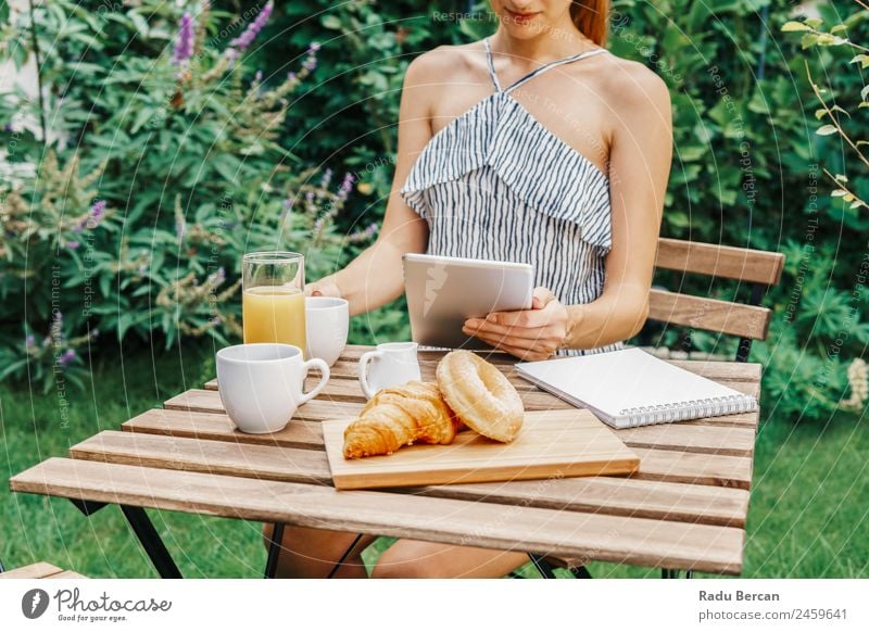 Junge und attraktive Frau beim Frühstück am Morgen im grünen Garten mit französischem Croissant, Donuts, Kaffeetasse, Orangensaft, Tablette und Notizbuch auf Holztisch.