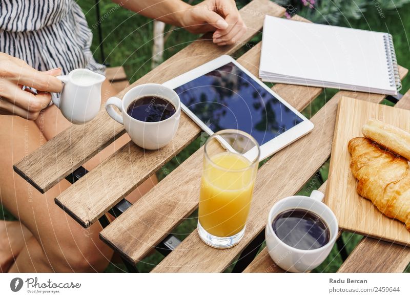 Junge und attraktive Frau beim Frühstück am Morgen im grünen Garten mit französischem Croissant, Donuts, Kaffeetasse, Orangensaft, Tablette und Notizbuch auf Holztisch.