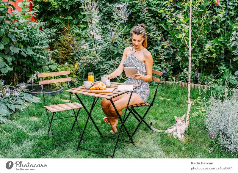 Junge und attraktive Frau beim Frühstück am Morgen im grünen Garten mit französischem Croissant, Donuts, Kaffeetasse, Orangensaft, Tablette und Notizbuch auf Holztisch.