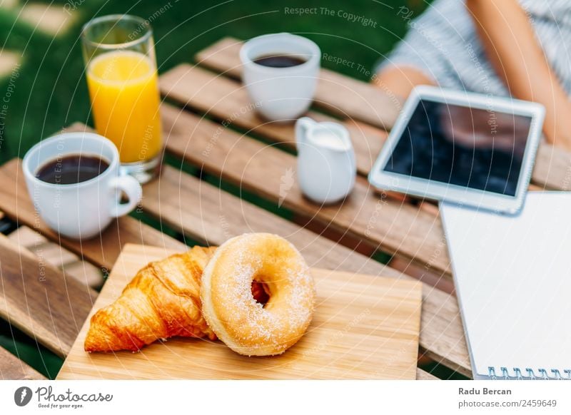 Junge und attraktive Frau beim Frühstück am Morgen im grünen Garten mit französischem Croissant, Donuts, Kaffeetasse, Orangensaft, Tablette und Notizbuch auf Holztisch.