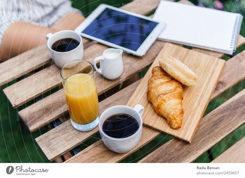 Junge und attraktive Frau beim Frühstück am Morgen im grünen Garten mit französischem Croissant, Donuts, Kaffeetasse, Orangensaft, Tablette und Notizbuch auf Holztisch.