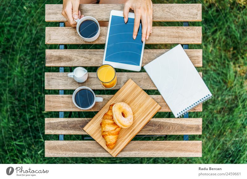 Junge und attraktive Frau beim Frühstück am Morgen im grünen Garten mit französischem Croissant, Donuts, Kaffeetasse, Orangensaft, Tablette und Notizbuch auf Holztisch.