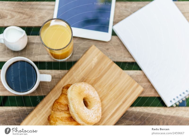 Frühstück im Grünen Garten mit französischem Croissant, Donuts, Kaffeetasse, Orangensaft, Tablette und Notizen Buch auf Holztisch Tisch Hintergrundbild Morgen