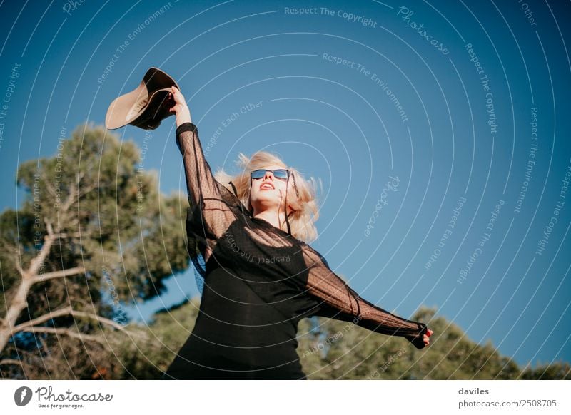 Glückliche blonde Frau, die bei Sonnenuntergang im Wald den Arm mit einem Hut in der Hand hebt. Sonnenbrille Völker Natur Kaukasier Ferien & Urlaub & Reisen