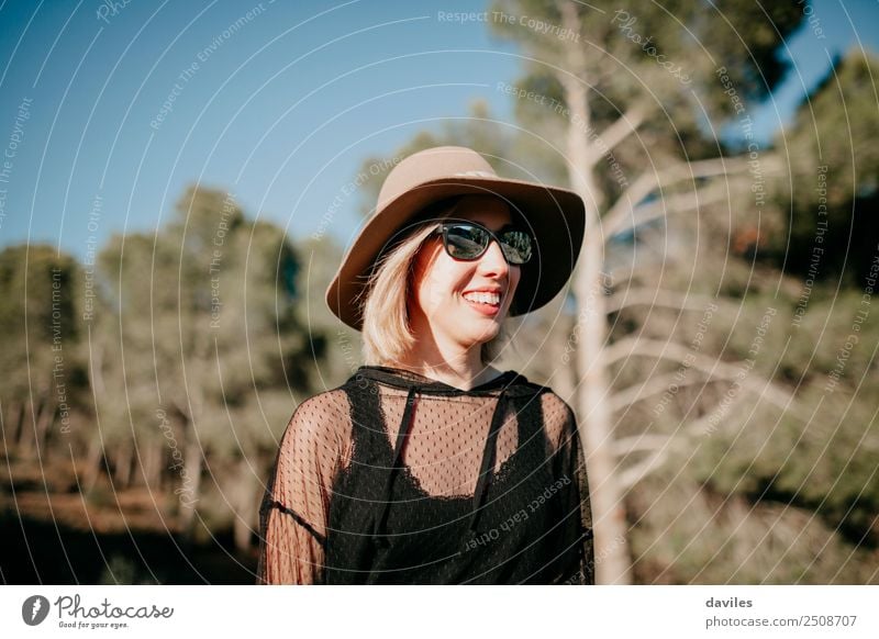 Hübsches blondes Mädchen mit Sonnenbrille und Hut, das sich in der Natur mit einem Kiefernwald im Hintergrund amüsiert Lifestyle Stil Freude Erholung