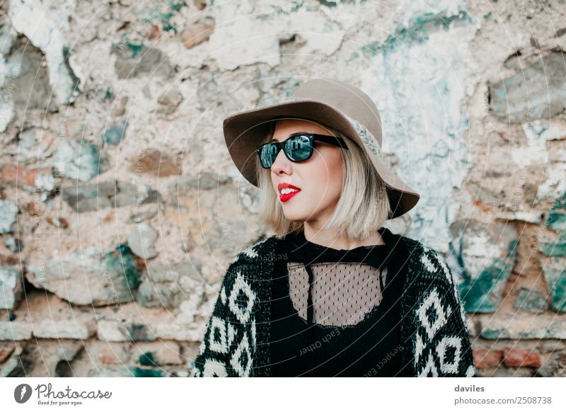 Bildnis eines blonden Mädchens mit Hut und Sonnenbrille, das mit einer Steinmauer im Hintergrund wegschaut. Lifestyle kaufen Reichtum elegant Stil Freude schön