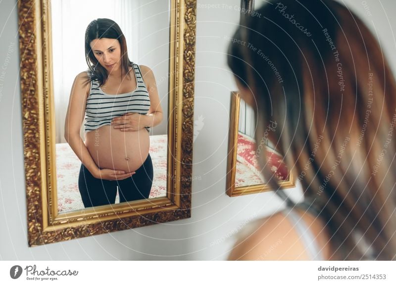 Schwangere Frau, die ihren Bauch ansieht. Lifestyle schön Leben Spiegel Schlafzimmer Mensch Baby Erwachsene Eltern Mutter Hand Denken Wachstum warten