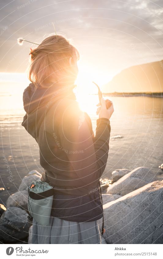 Gegenlichtaufnahme einer jungen Frau mit Pfeife und Blume im Haar im Porträt in der Rückansicht in der Mitternachtssonne am Strand eines Fjords Freiheit