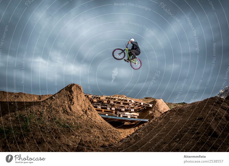 Junger Mann, der über ein Loch im Dirtjump-Kreislauf springt. Freude Erholung Berge u. Gebirge Sport Fahrradfahren Erwachsene Natur Erde Wolken Park Hügel