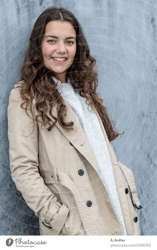 Junge Frau im Mantel Mensch Platz Mauer Wand Mode Bekleidung Stoff brünett langhaarig Locken Lächeln Coolness Freundlichkeit Fröhlichkeit schön blau braun grau