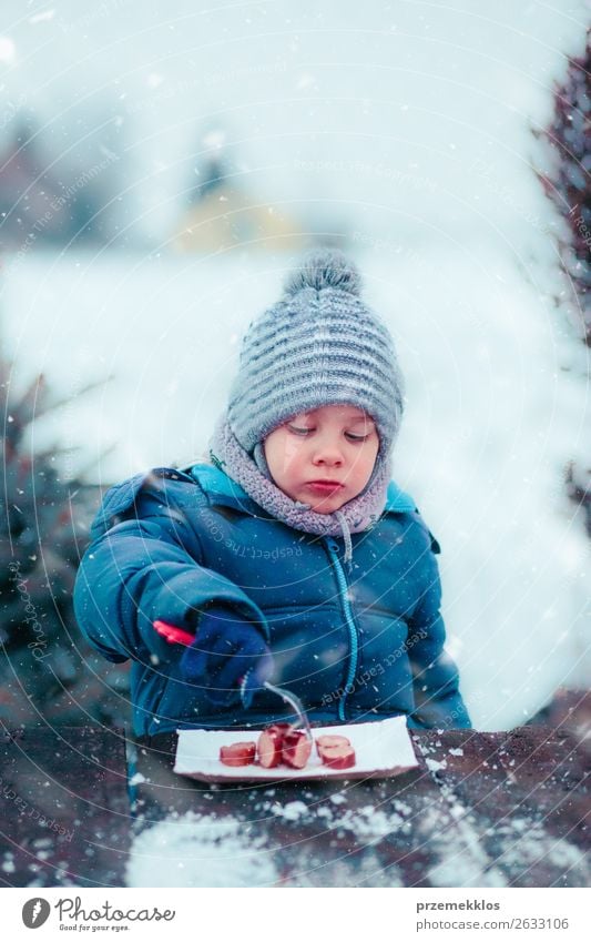 Junge isst gebratene Wurst beim Picknick im Winter im Freien. Wurstwaren Essen Lifestyle Freude Schnee Winterurlaub 3-8 Jahre Kind Kindheit Schneefall Mütze