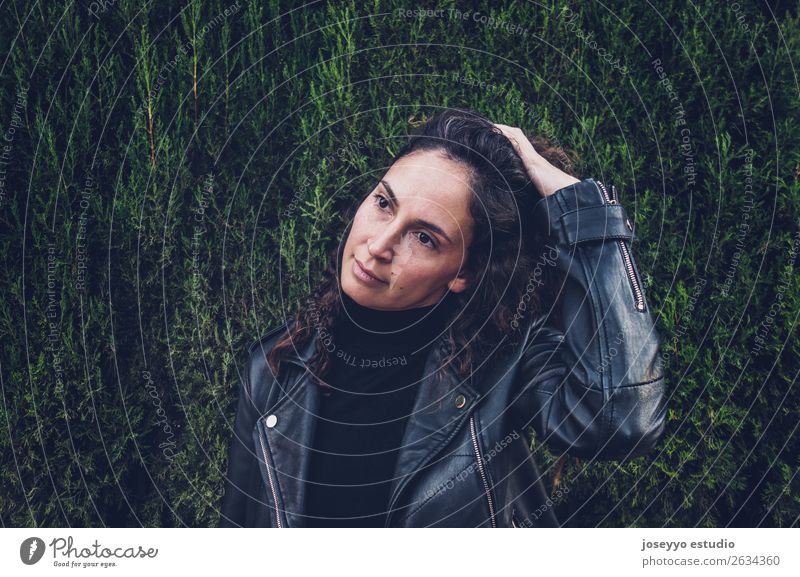 Die Frau trägt eine Lederjacke gegen eine Zypressenwand. Lifestyle Glück schön Gesicht Mensch Natur Pflanze Herbst Park Mode Jacke brünett berühren Lächeln