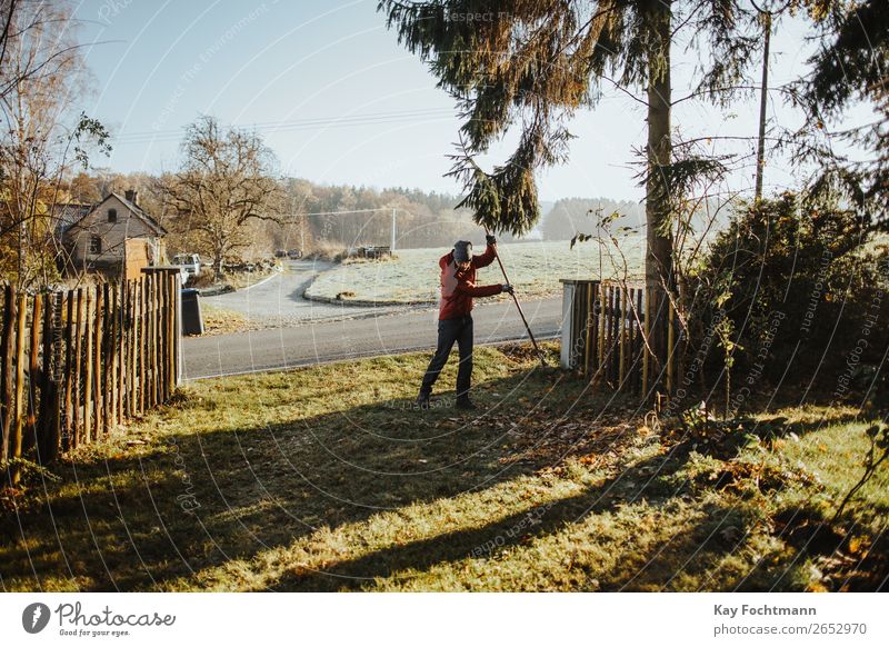 Kaukasischer Mann mit roter Jacke harkt im Herbst Blätter Aktivität Hinterhof Beanie Botanik Reinigen Säuberung fallen Fallout Laubwerk Garten Gärtner