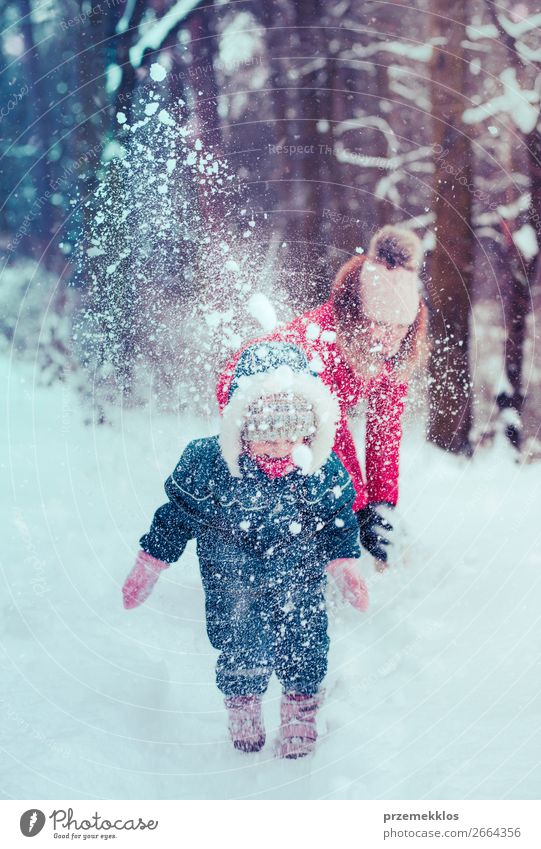 Mutter spielt mit ihrer kleinen Tochter im Freien an einem winterlichen Tag. Frau wirft Schnee auf ihr Kind. Familie verbringt Zeit zusammen und genießt die Winterzeit. Frau trägt roten Mantel und Wollmütze, Kleinkind trägt dunkelblauen Schneeanzug