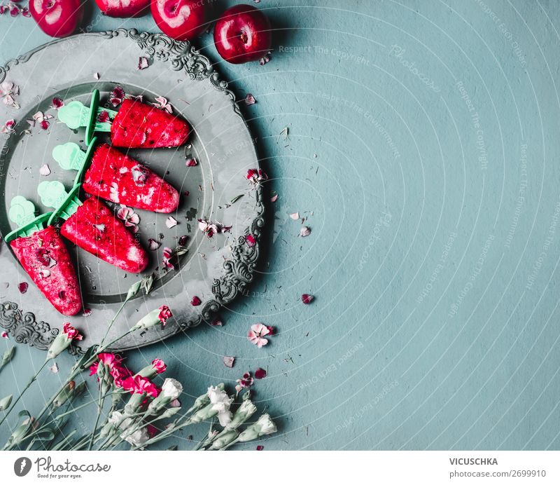 Obst und Beeren Eis am Stiel . Hausgemachtes Eis im Vintage-Teller auf blauem Küchentisch im Hintergrund mit Sommerblumen, Ansicht von oben. Gesunde Sommerdesserts. Gefrorene Säfte am Stiel. Veganes Eis